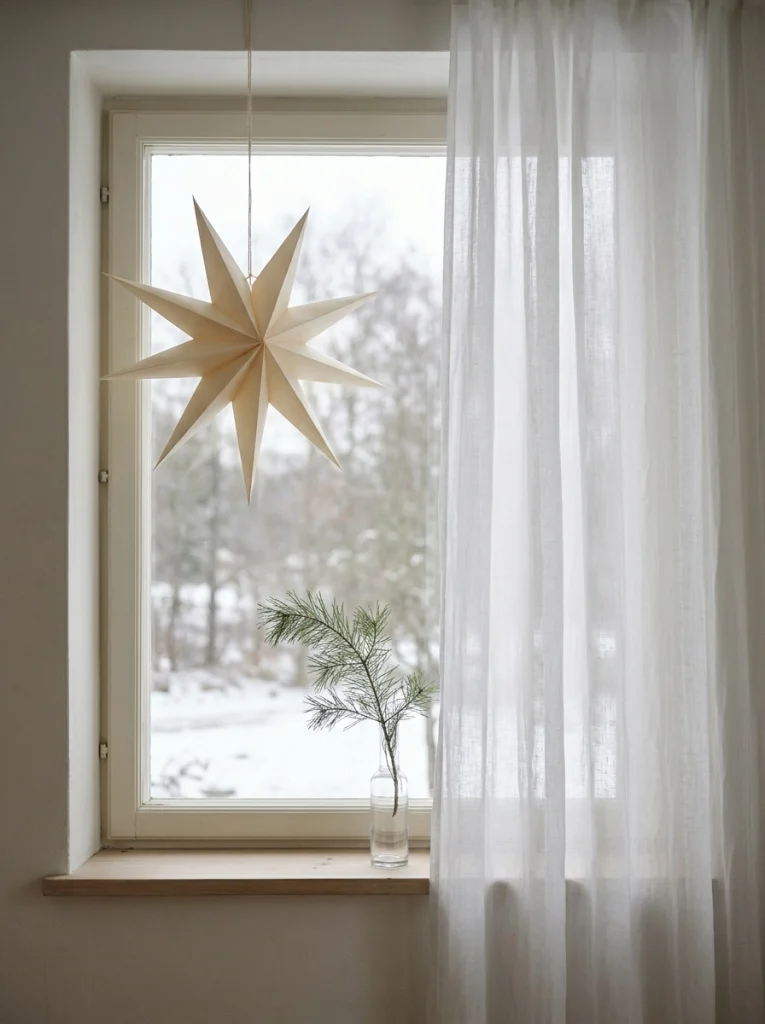 Minimalist Christmas window decor with a paper star, sheer curtains, and an evergreen branch.